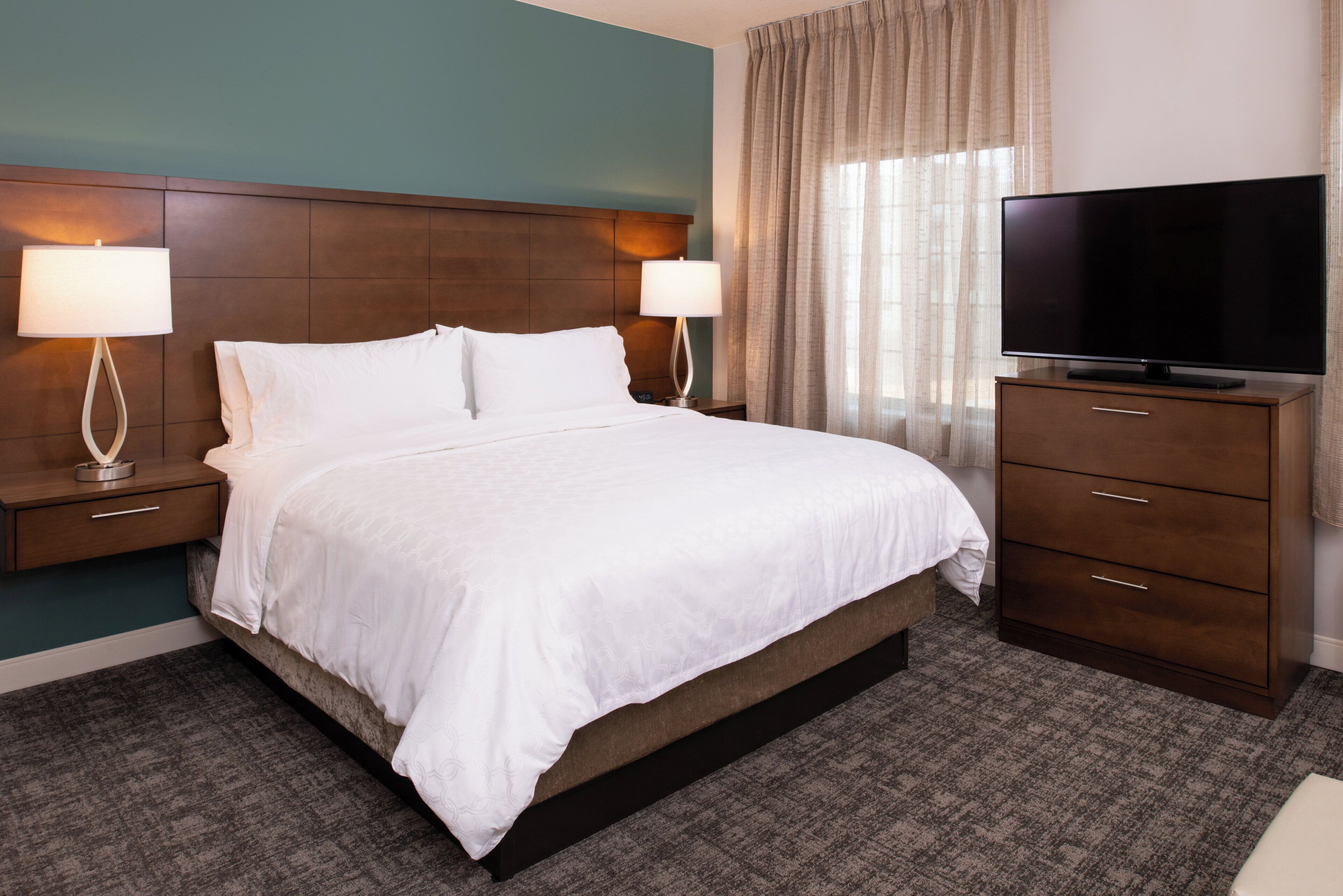 Habitación de hotel con una cama grande, dos mesitas de noche con lámparas, un televisor sobre un mueble de madera y vistas al exterior a través de cortinas.