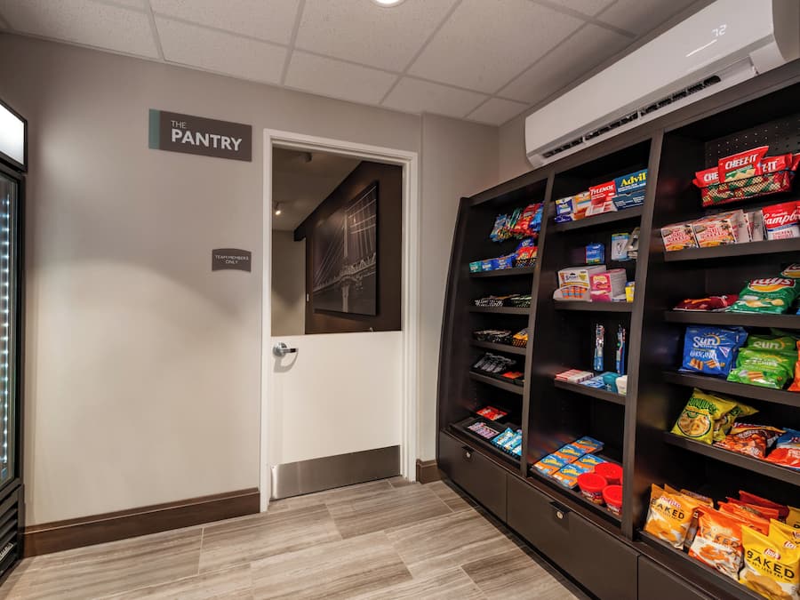 Ein Vorratsschränkchen mit einem Kühlschrank, der mit Getränken und Snacks gefüllt ist, eine Tür mit der Aufschrift 'TEAM MEMBERS ONLY' und Regale mit verschiedenen Lebensmitteln.