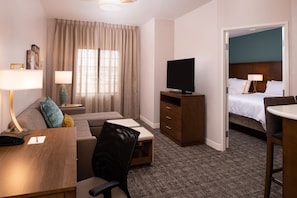 Relax in a comfortable seating area with a sofa and chair, featuring a flat-screen TV and a neatly made bed with white linens.
