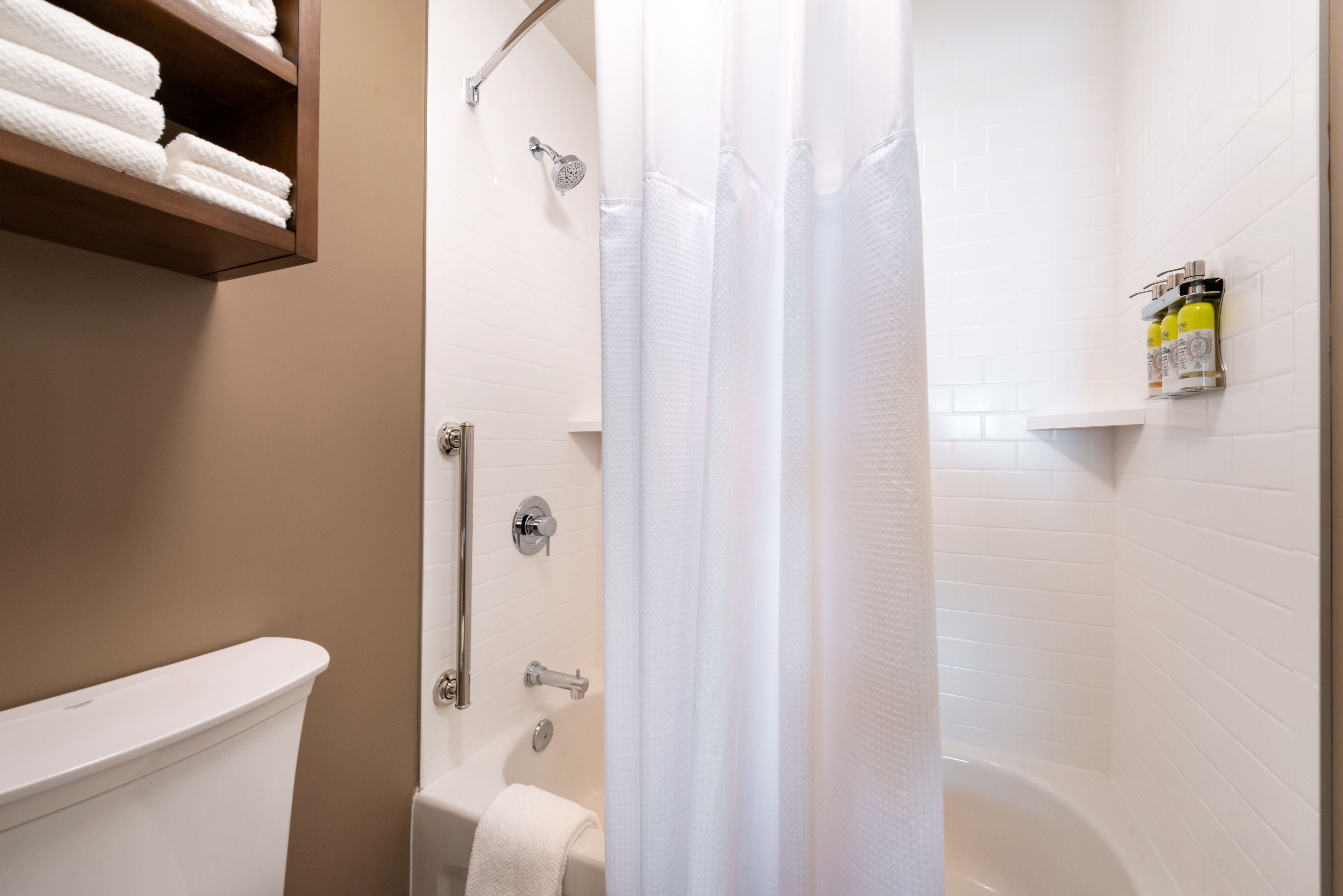 A bathroom with a toilet, shower, and bathtub. There is a shelf with towels and a bottle of shampoo.