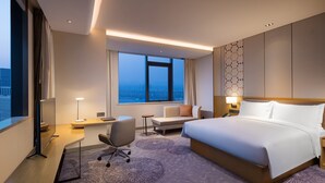 A hotel room with a large bed, a desk, a chair, and a view of the city through a large window. - Holiday Inn Tianjin Xiqing by IHG (Tianjin)