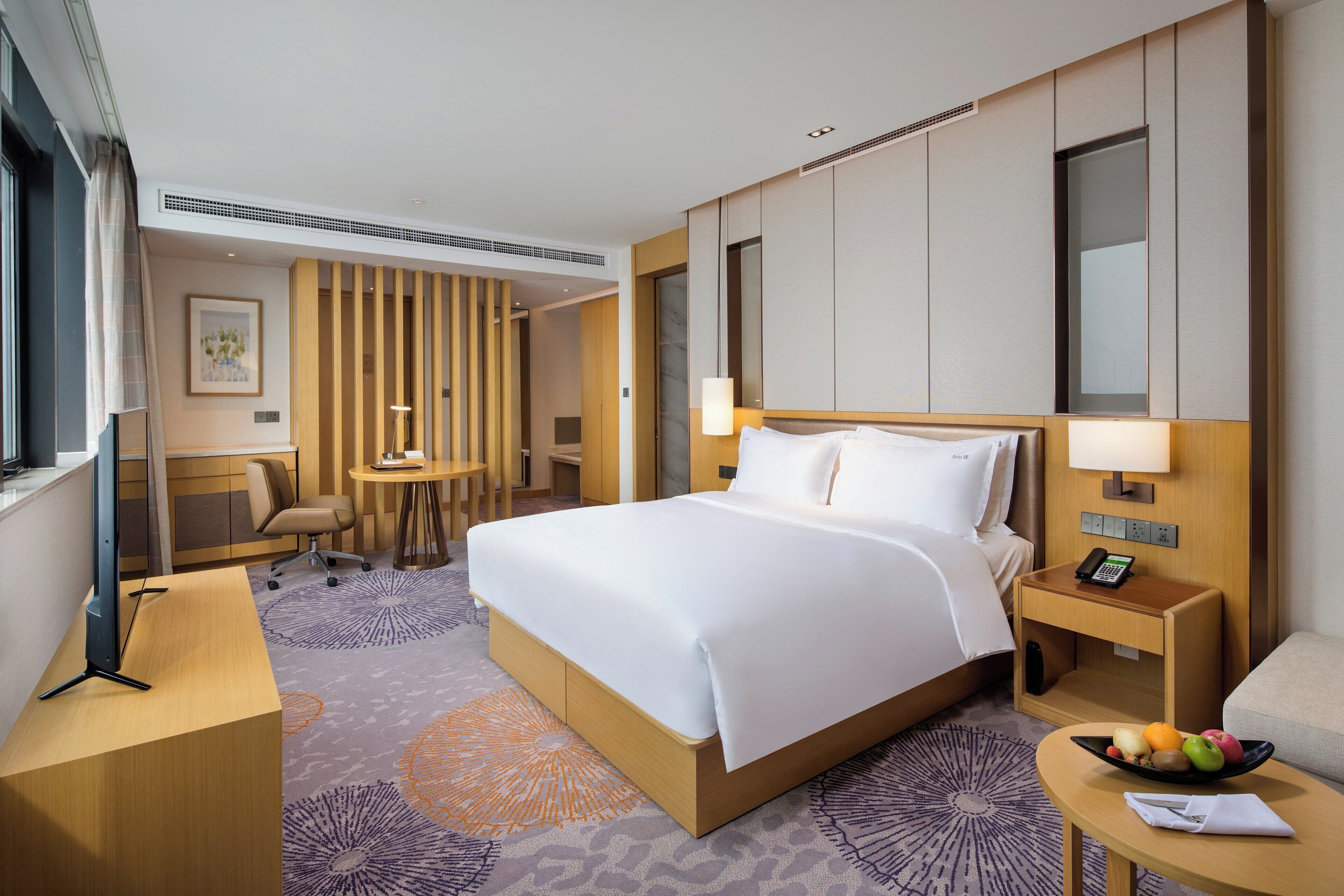 A hotel room with a large bed, a desk, a chair, a small table, a mirror, and a fruit bowl.