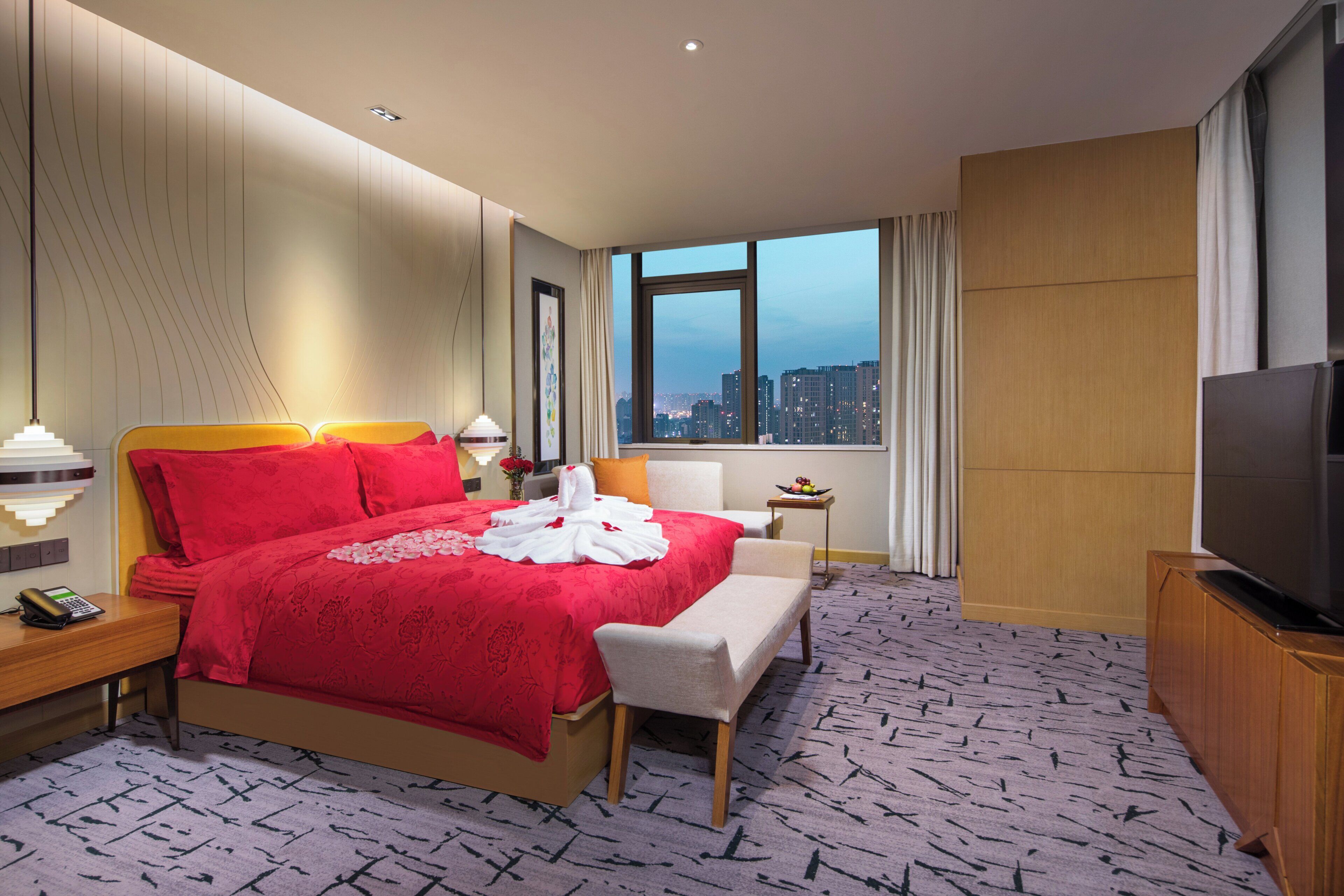 A hotel room with a large bed, a television, a desk with a telephone, and a window with a city view.
