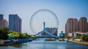 A Ferris wheel with a blue structure in the foreground, a river, and high-rise buildings in the background. - Holiday Inn Tianjin Xiqing by IHG (Tianjin)