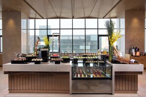 A buffet setup with various food items and a glass display case, set against a backdrop of large windows with a cityscape view. - Holiday Inn Tianjin Xiqing by IHG (Tianjin)