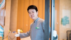 A person in a grey uniform standing in a hotel lobby with a name tag, a glass partition, and a wooden wall in the background. - Holiday Inn Tianjin Xiqing by IHG (Tianjin)