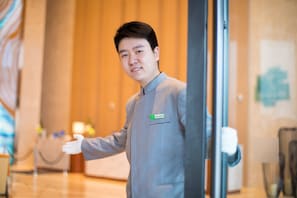 A person in a grey uniform standing in a hotel lobby with a name tag, a glass partition, and a wooden wall in the background. - Holiday Inn Tianjin Xiqing by IHG (Tianjin)