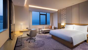 A hotel room with a large bed, a desk, a chair, a small table, a window with a view, and a patterned carpet.