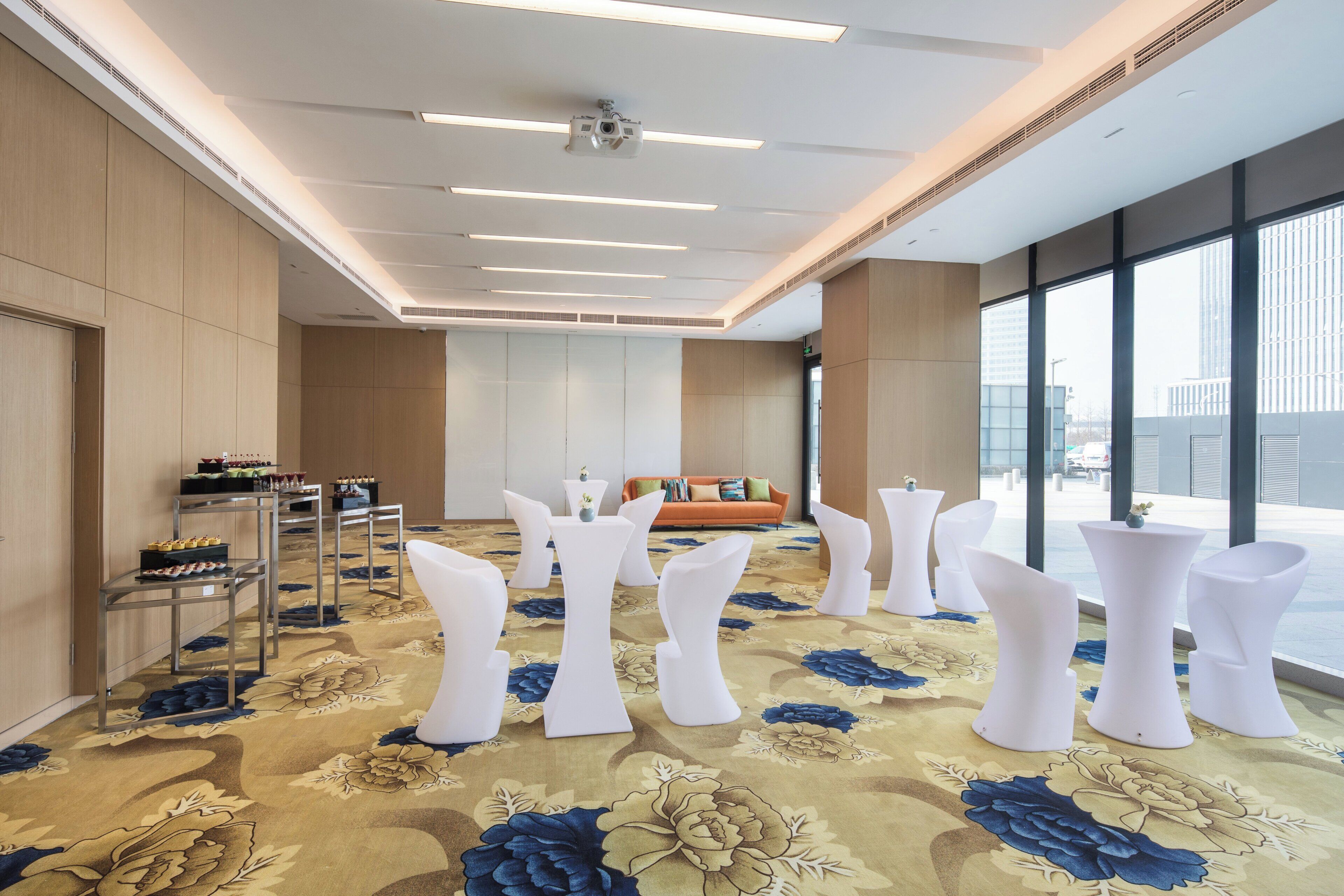 Una sala de conferencias moderna con techo alto, proyector, sofá y alfombra estampada con motivos florales.