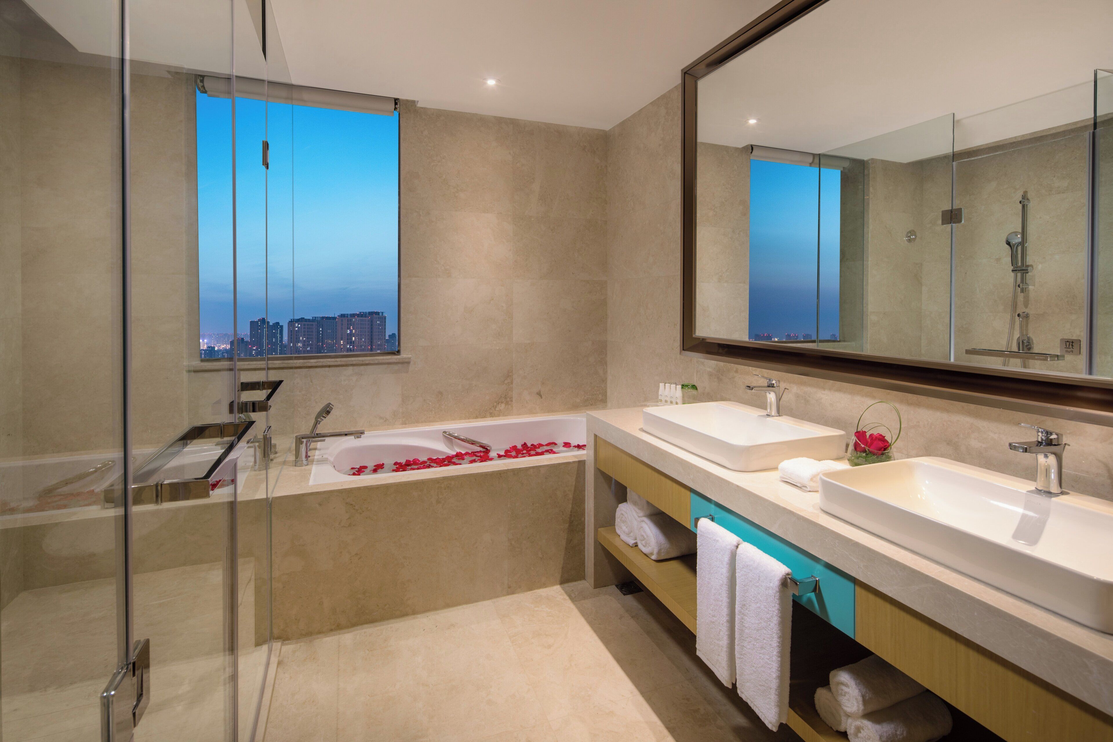 Un baño moderno con cabina de ducha de vidrio, una bañera con pétalos de rosa y vistas a la ciudad a través de una ventana.