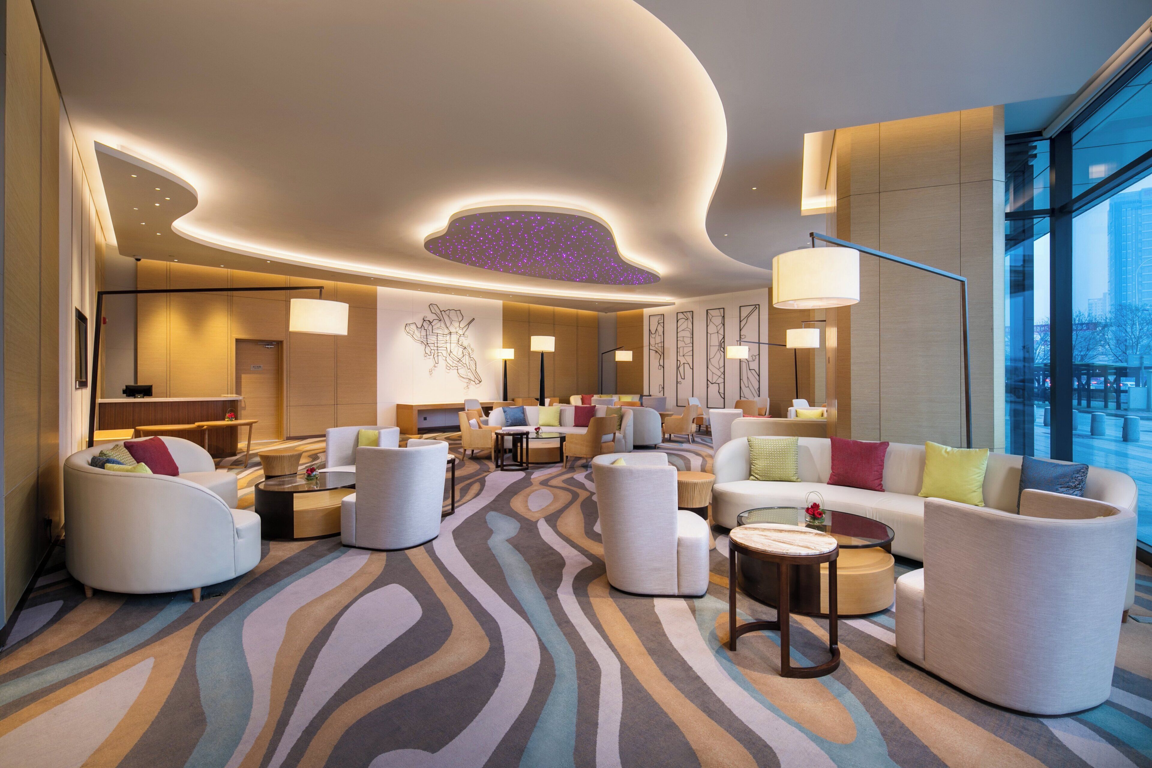 A modern hotel lobby with a central seating area, a large window offering a city view, and a ceiling with a unique light feature.