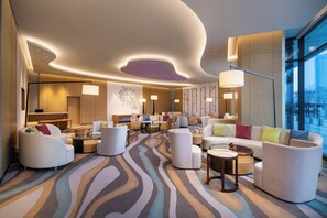A modern hotel lobby with a central seating area, a large window offering a city view, and a ceiling with a unique light feature.