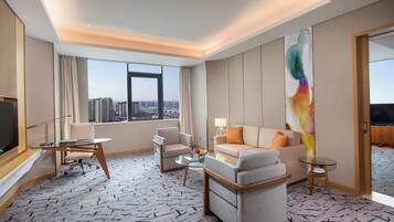 A hotel room with a large window, a sofa, a chair, a small table, and a TV.