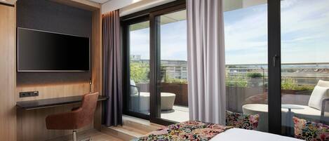 Una habitación de hotel moderna con una cama grande, un escritorio con televisor de pantalla plana, un sillón cómodo y un balcón con mobiliario exterior.