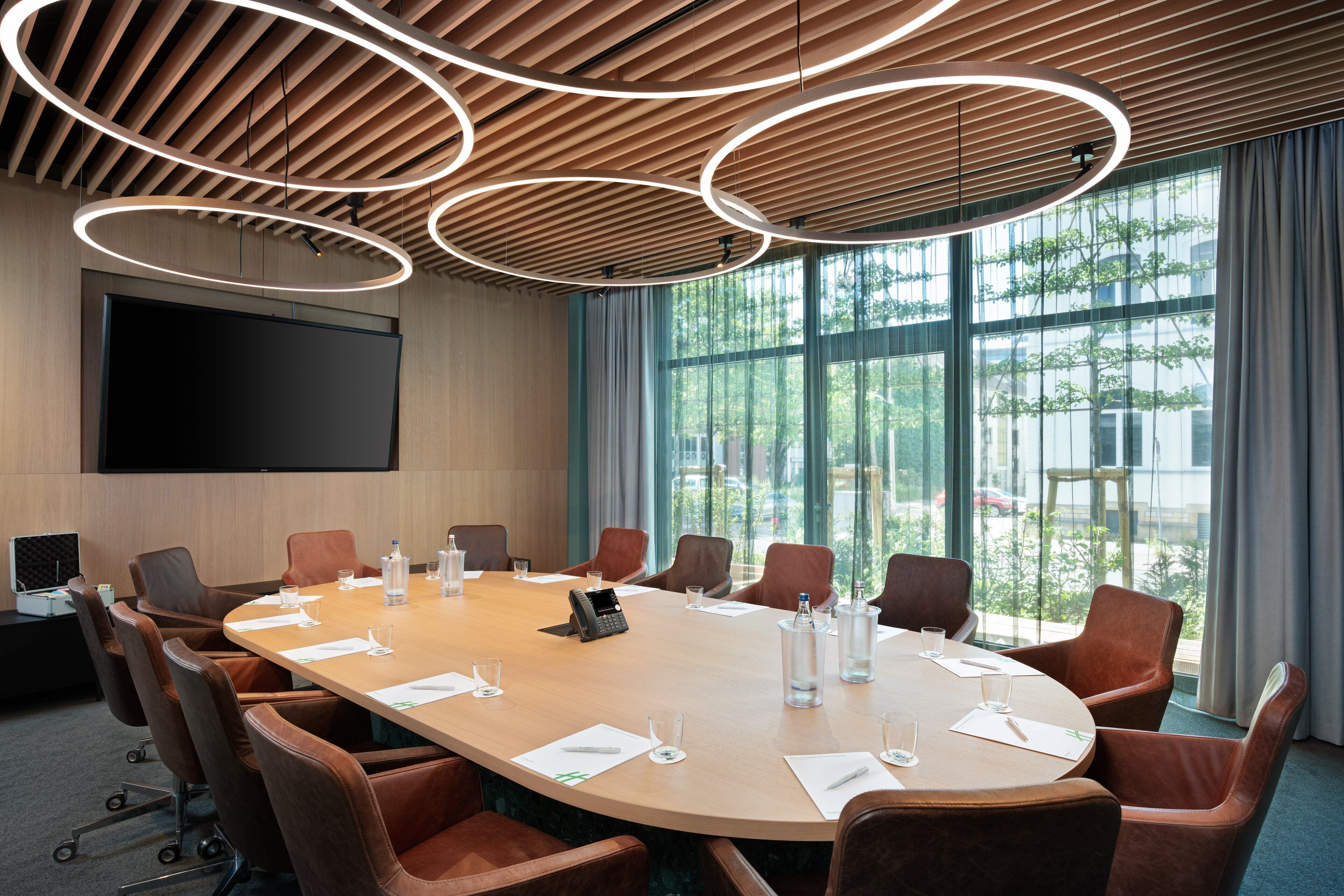 A conference room with a large oval table, leather chairs, a flat-screen TV, and a view of trees through large windows.