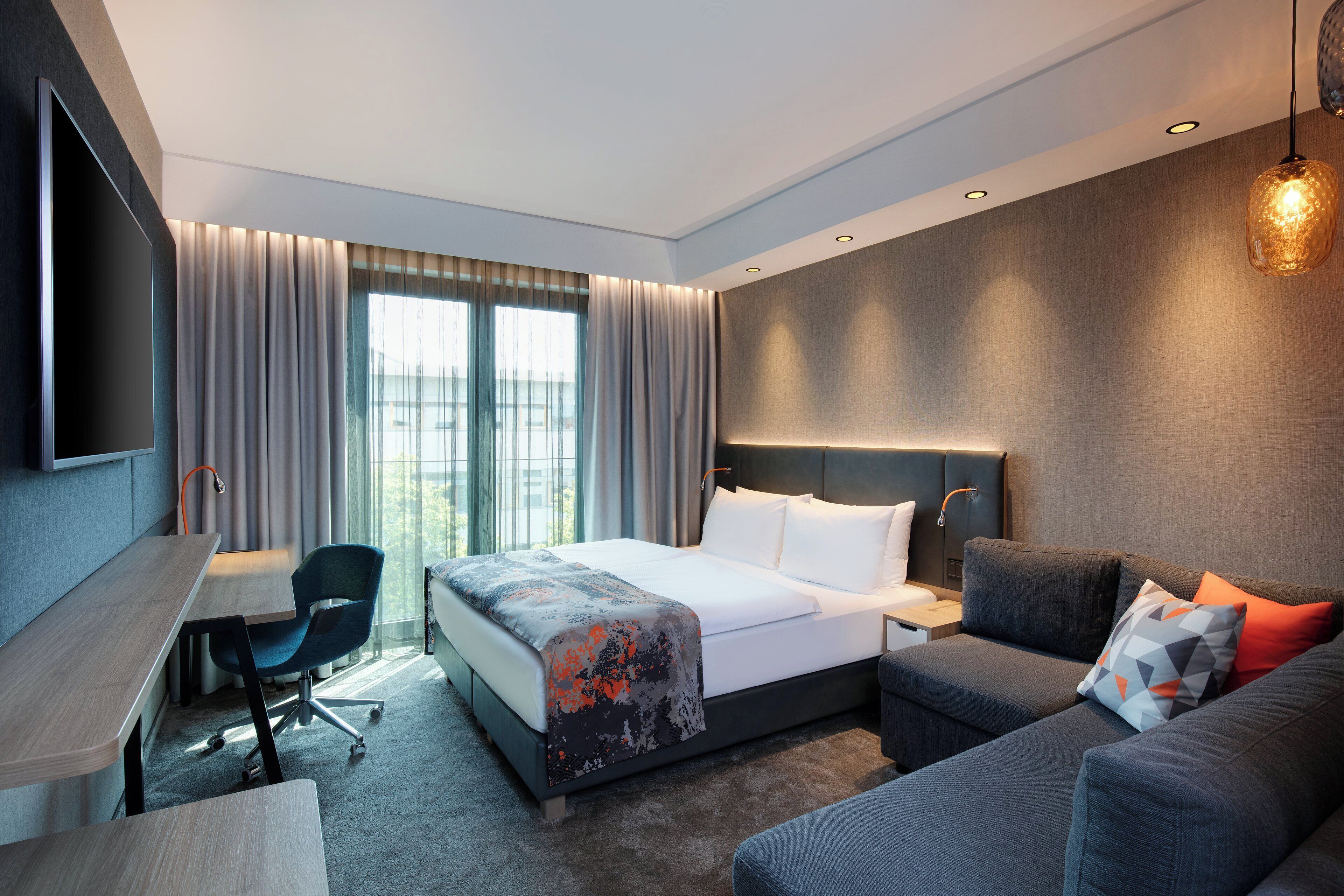 Ein modernes Hotelzimmer mit Bett, Schreibtisch, Stuhl, Sofa und einem an der Wand befestigten Fernseher.