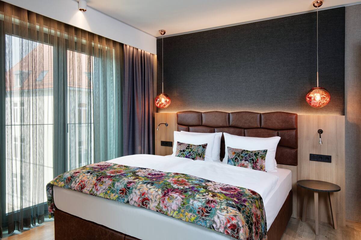 A modern hotel room with a large bed, a headboard with a built-in shelf, two bedside tables, and a large window with curtains.