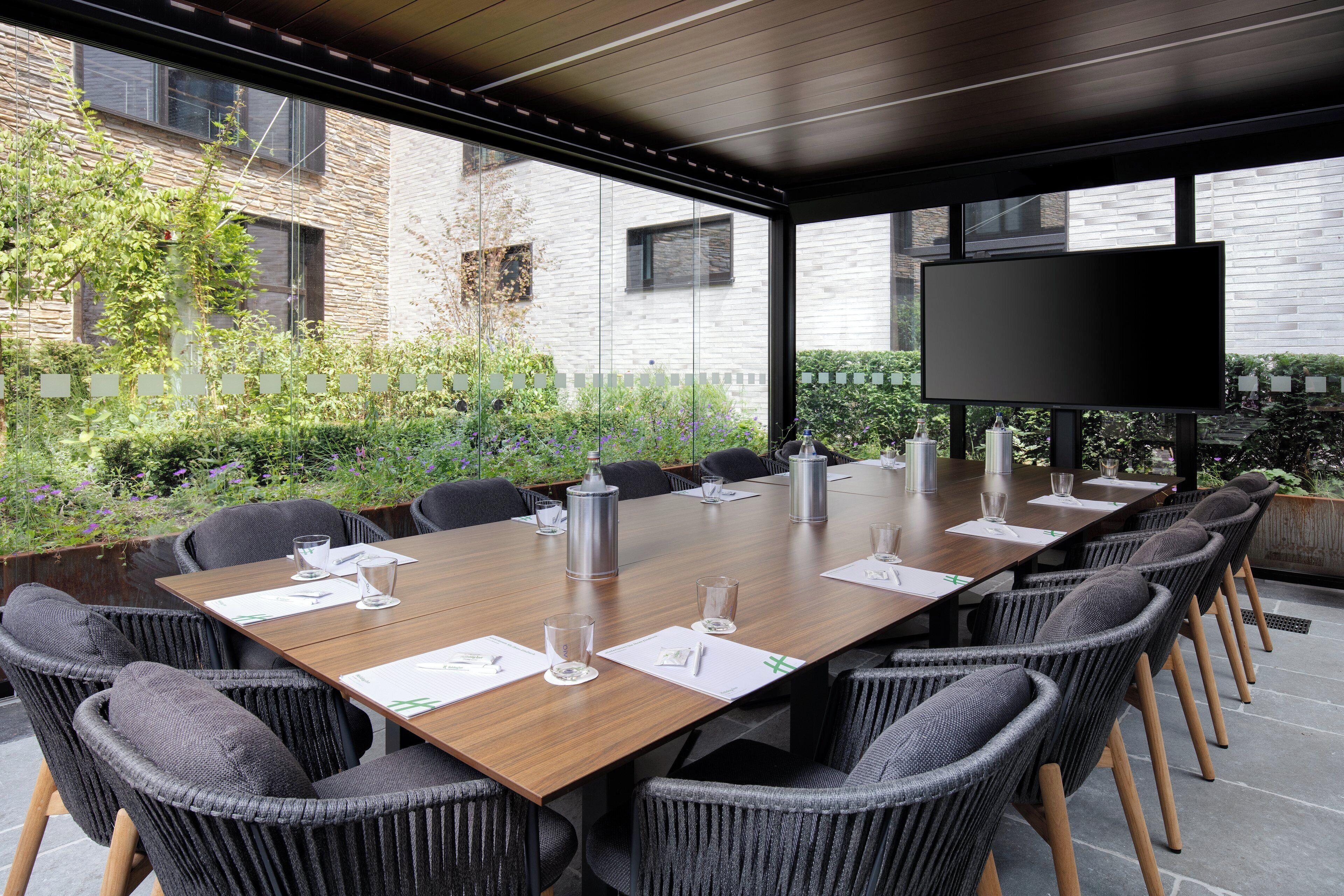 Une salle de réunion extérieure dotée d’une longue table, de chaises, d’un téléviseur à écran plat et offrant une vue sur un bâtiment en briques.