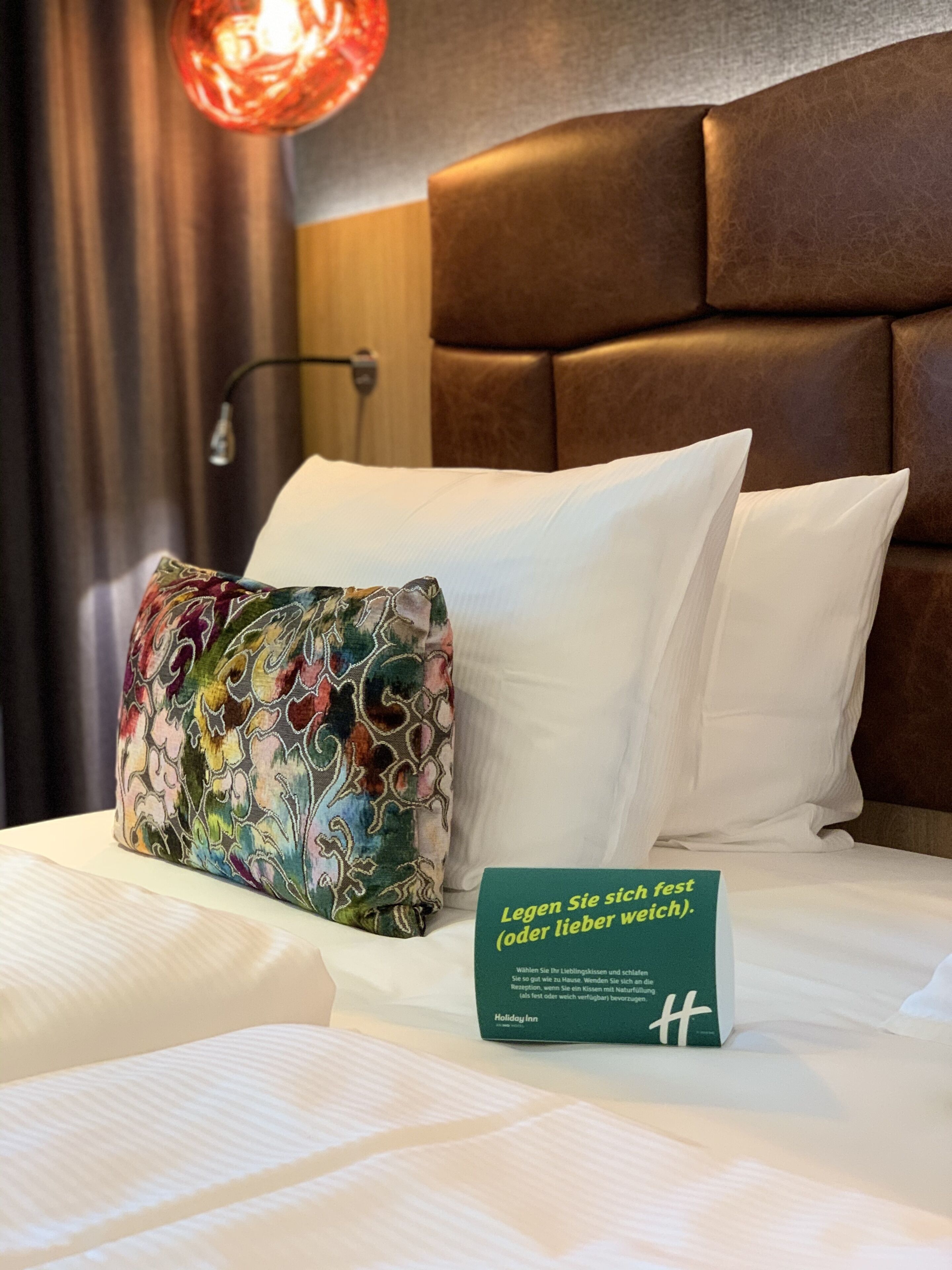 A hotel room with a bed, a colorful pillow, a white pillow, and a sign with text in German.