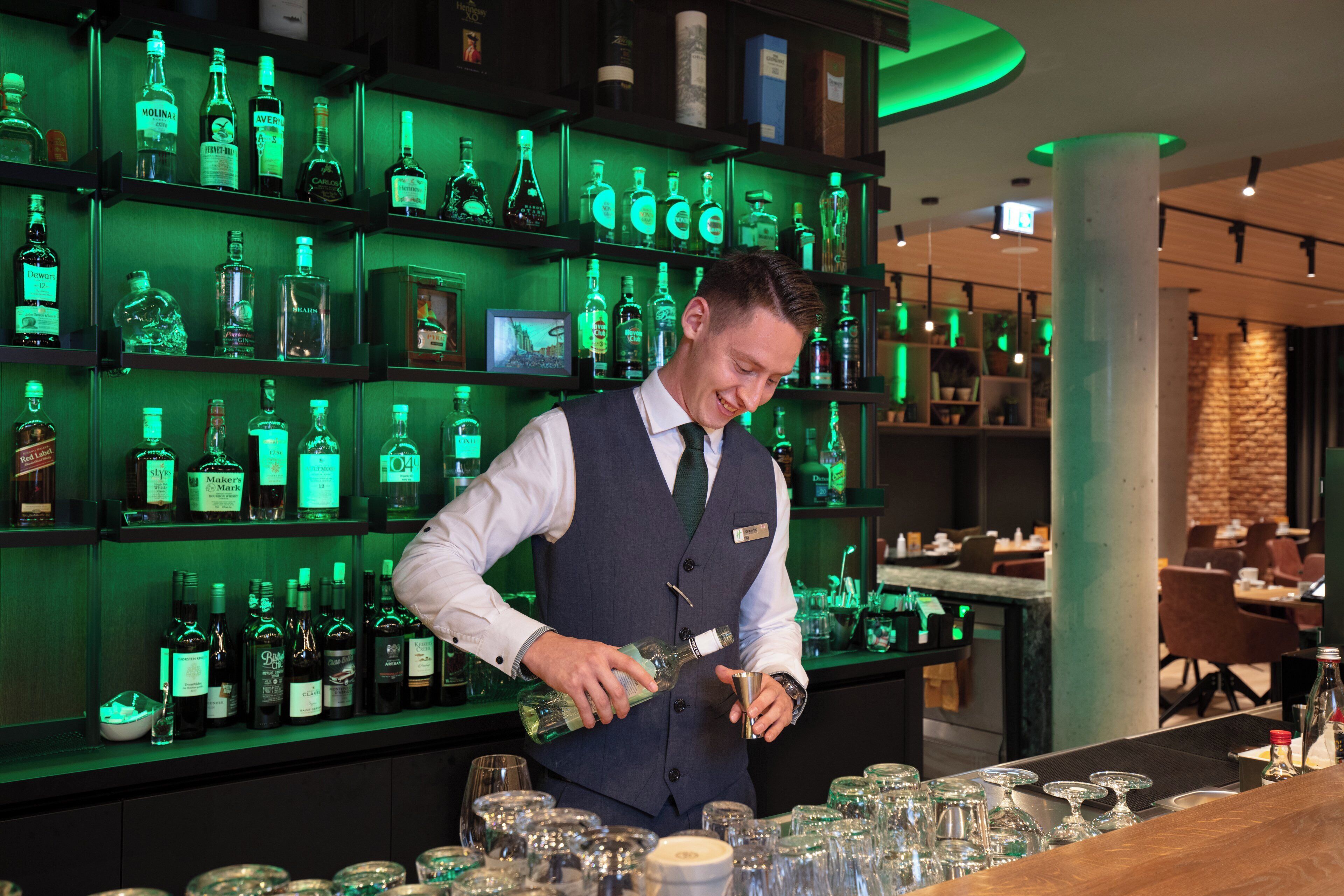 A bartender preparing drinks at a bar with shelves of liquor bottles in the background.