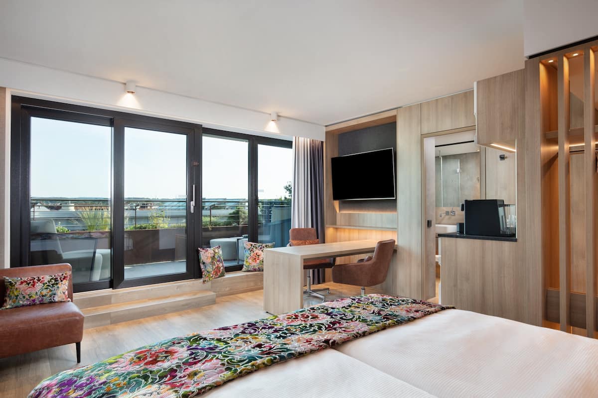 A modern hotel room with a large bed, a desk, and a chair. There is a TV mounted on the wall and a balcony with a view of the city.