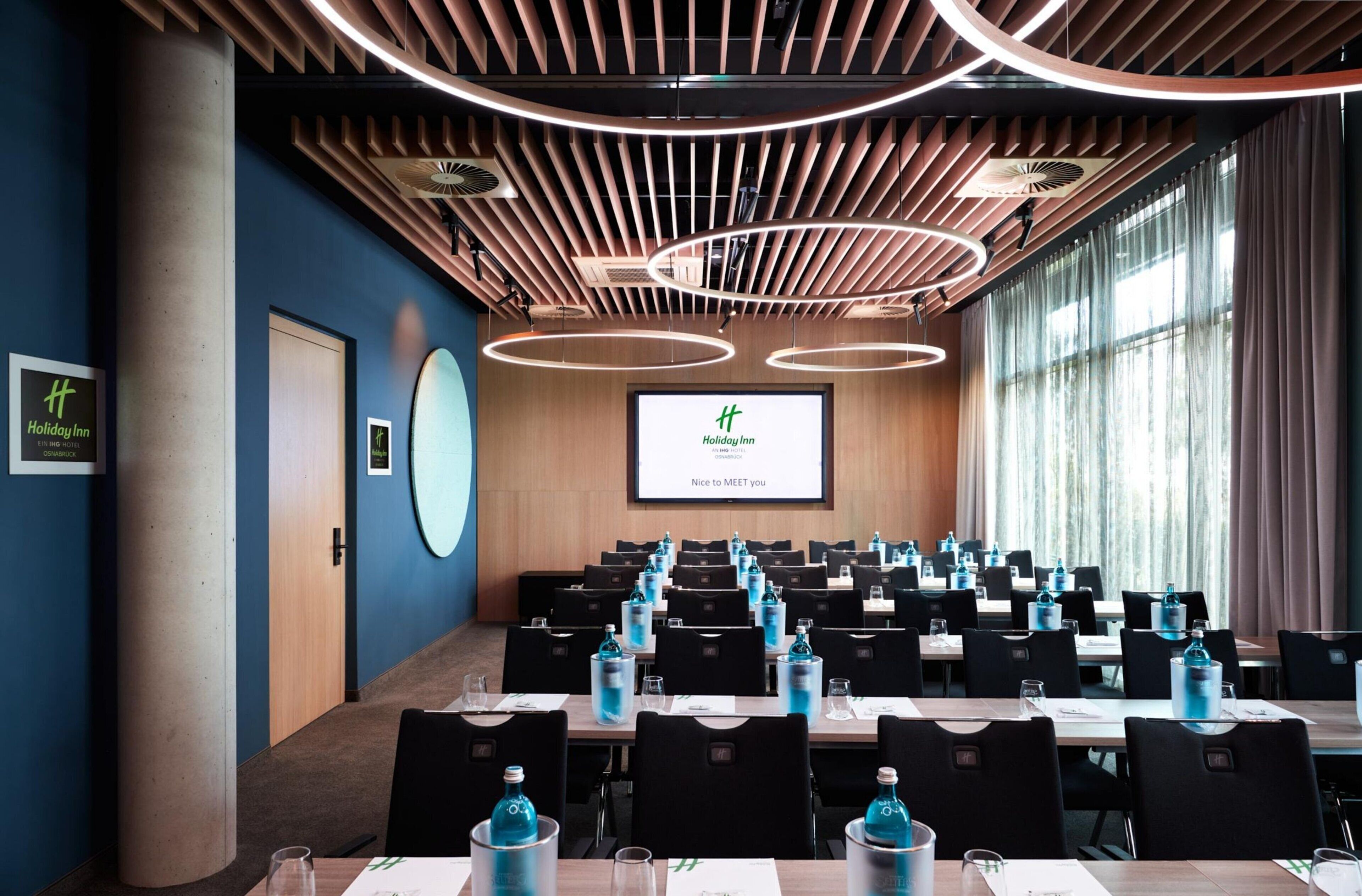 A conference room with a large screen displaying a welcome message, multiple tables with chairs, and a modern ceiling design.