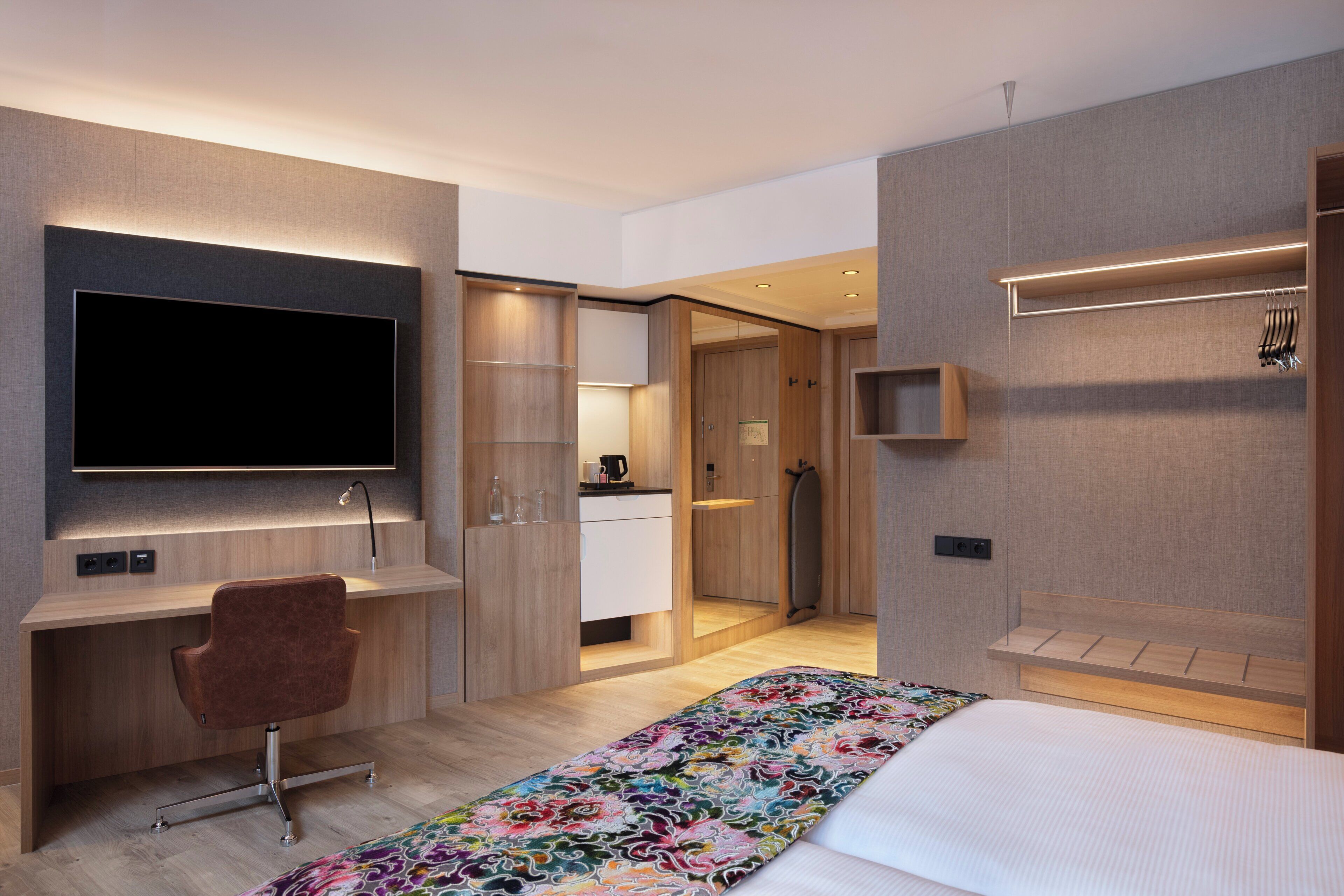 A modern hotel room with a flat-screen TV, a desk, a chair, a bed with a colorful bedspread, and a walk-in closet.