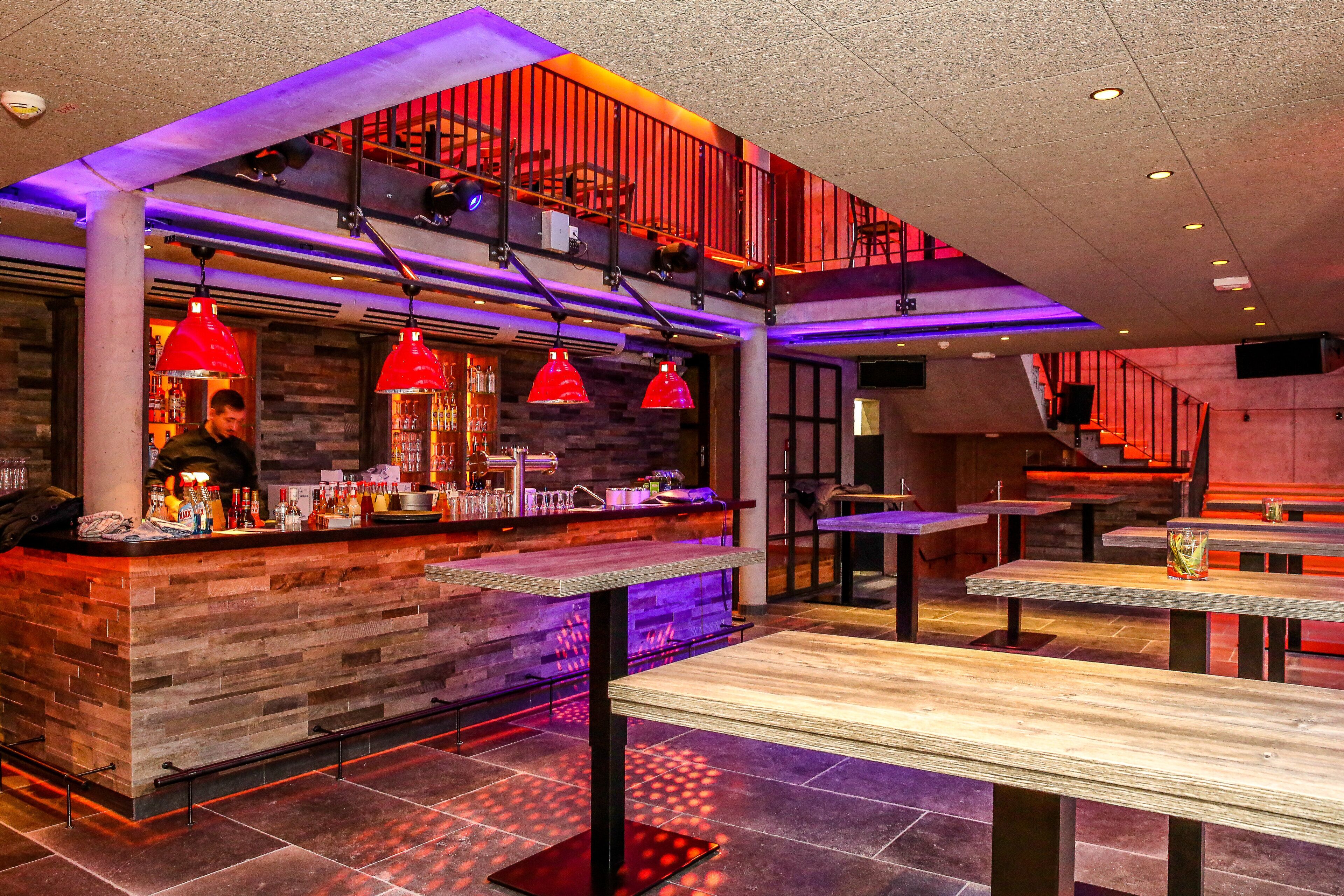 A modern bar area with a wooden counter, red pendant lights, and a staircase leading to an upper level.