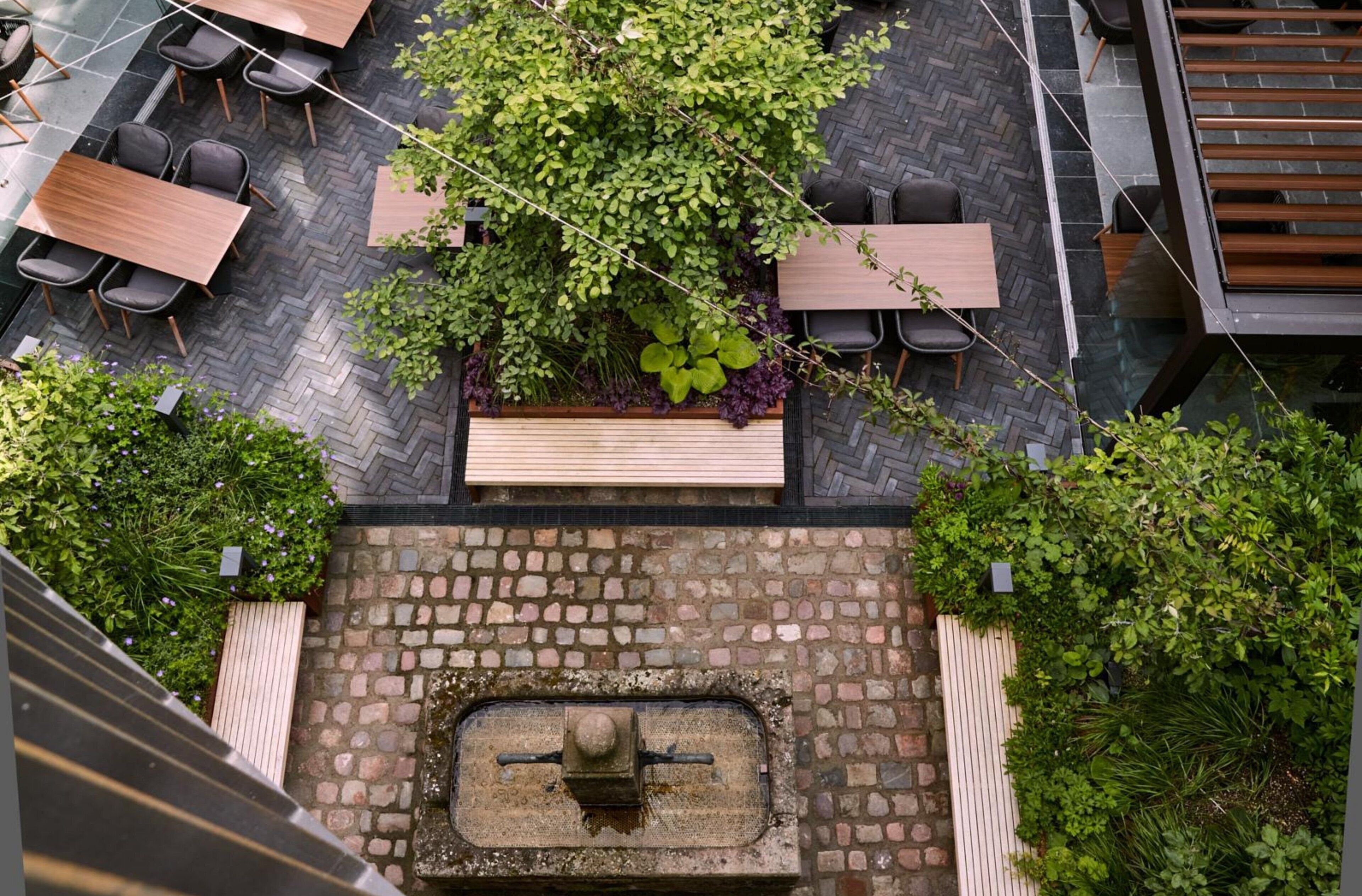 Un giardino pensile con tavoli e sedie in legno, immerso nel verde e con una fontana in pietra.