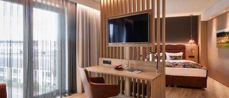 Una habitación de hotel moderna con un escritorio de madera, un televisor de pantalla plana grande, una cama con cabecera y un ventanal con cortinas.