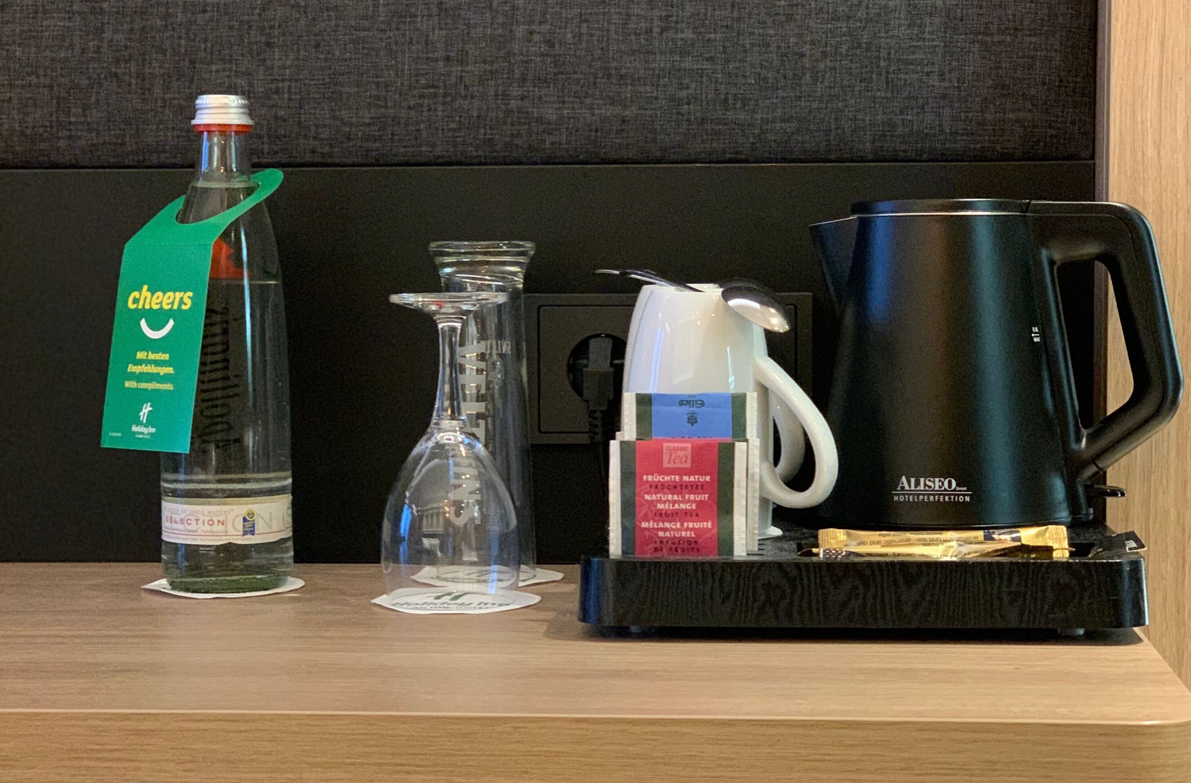Un mini-bar d’hôtel comprenant une bouteille d’eau, une carafe en verre, un distributeur de thé et une machine à café.