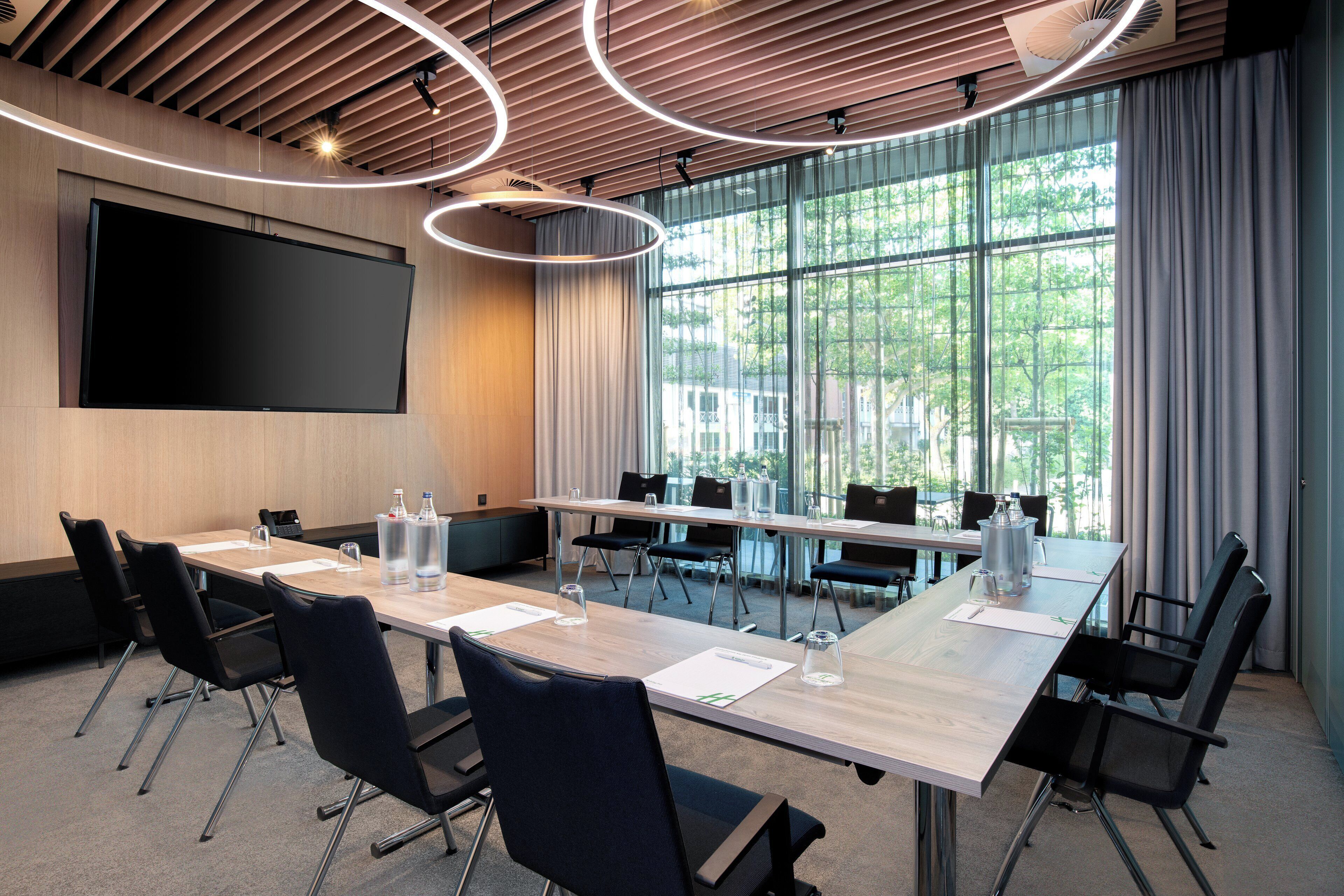 A conference room with a large table, chairs, a screen, and a view of greenery through large windows.