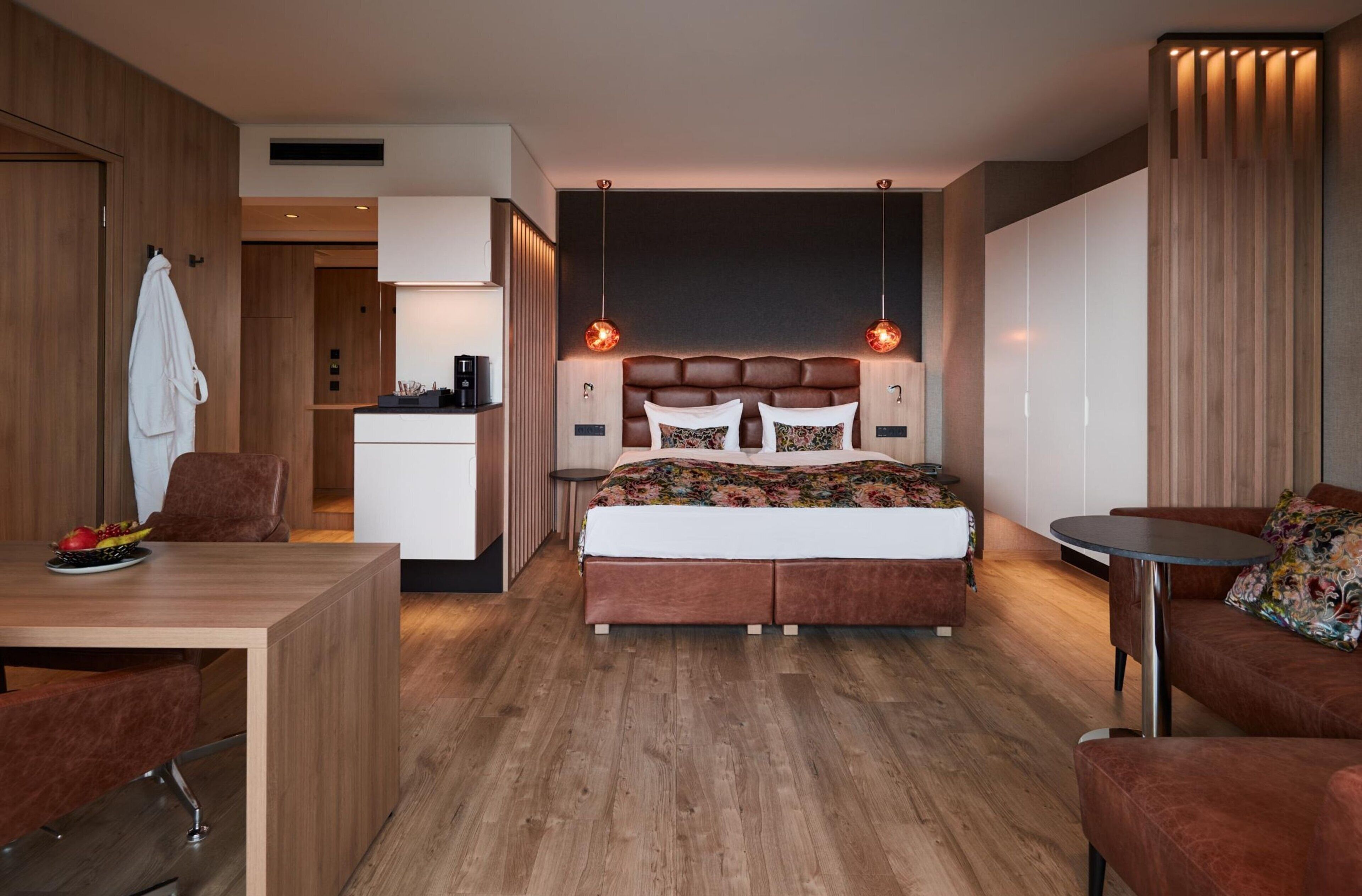 A modern hotel room with a wooden floor, a central bed with a headboard, a small desk, a round table, and a sofa.