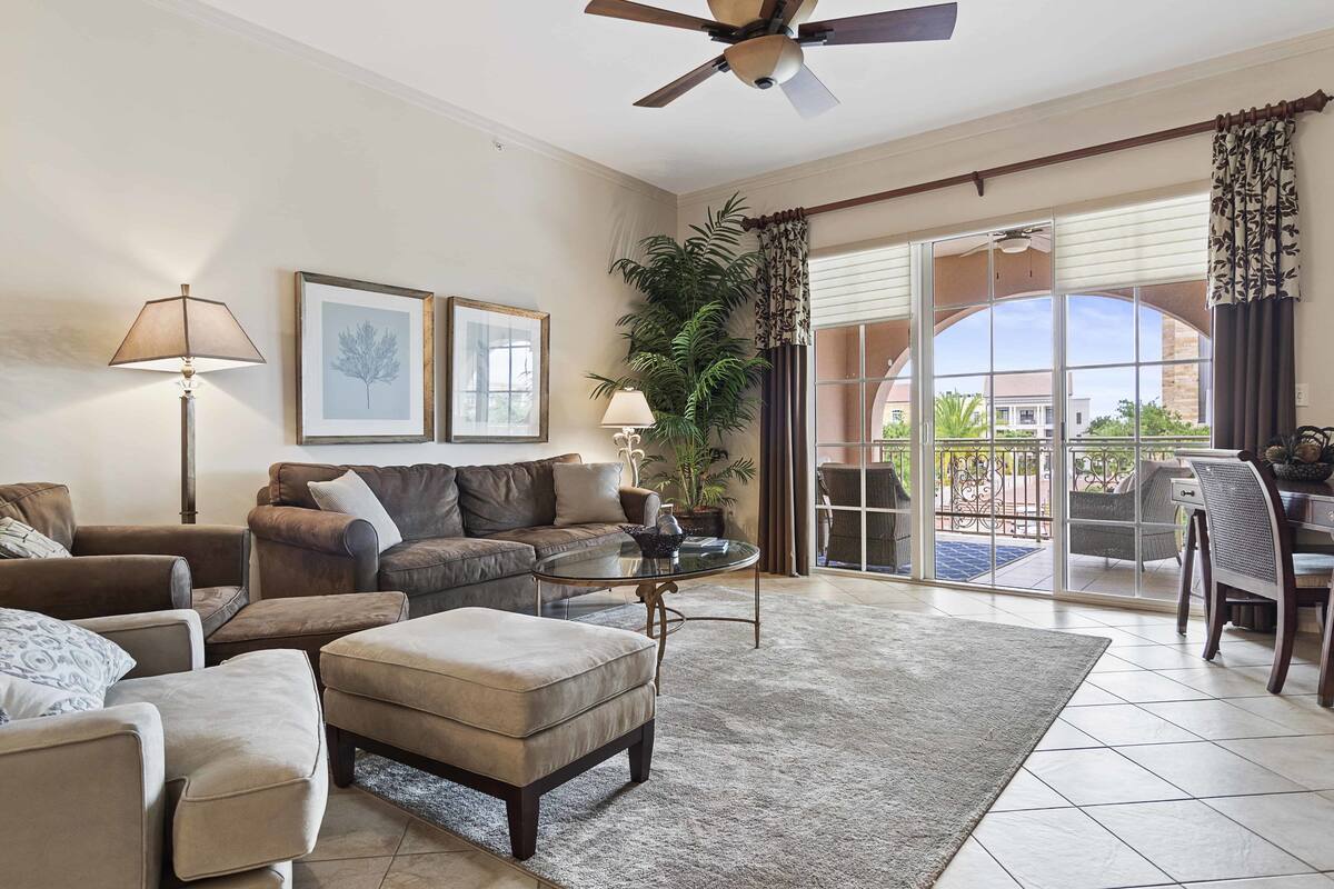 A spacious living room with a sofa, ottoman, and a glass coffee table. There is a ceiling fan, a potted plant, and a view of the outside through large sliding glass doors.