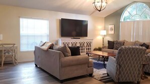 A living room with a sofa, armchairs, a coffee table, a TV, and a window with blinds.