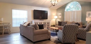 A living room with a sofa, armchairs, a coffee table, a TV, and a window with blinds.