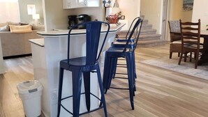 A kitchen with a bar area, a dining table with chairs, a trash bin, and a staircase leading to another room.
