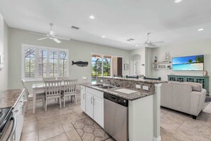 A modern kitchen with white cabinets, a granite countertop, and a dishwasher. There is a dining area with white chairs and a table. A large window with blinds is visible, and a TV is mounted on the wall.