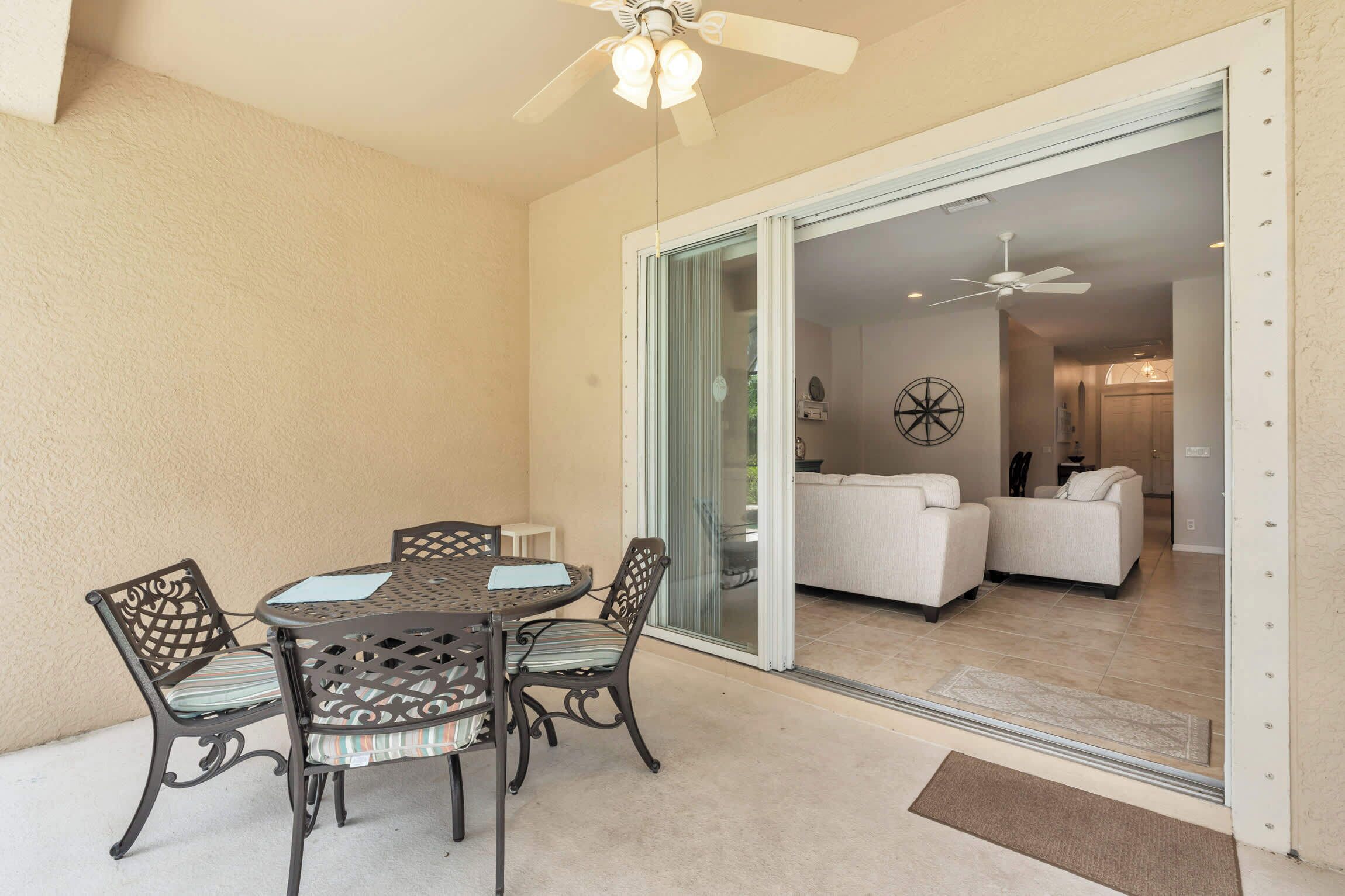 A patio with a round table and chairs, a ceiling fan, and a sliding glass door leading to a living area with a sofa and a decorative wall piece.