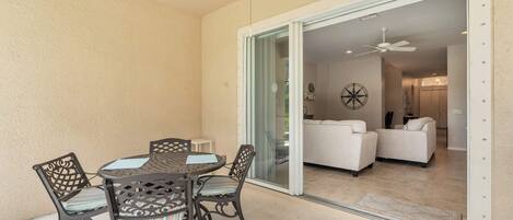 A patio with a round table and chairs, a ceiling fan, and a sliding glass door leading to a living area with a sofa and a decorative wall piece.