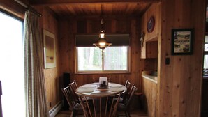 Comedor de madera con una mesa redonda, sillas, una lámpara colgante, un ventanal con cortinas y un cuadro en la pared.