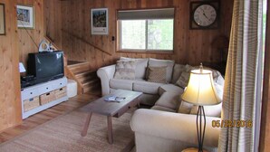 Interior de una cabaña de madera con sofá, mesa de centro, televisor y un reloj en la pared.