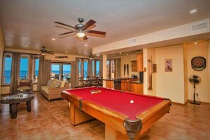A pool table in a spacious room with a view of the ocean, a ceiling fan, and a bar area.