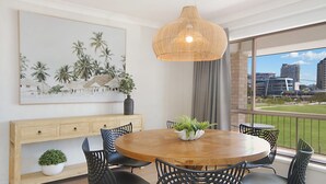 A dining area with a round wooden table, black chairs, a hanging wicker light fixture, a large framed tropical artwork, and a view of buildings and greenery through large windows. - Tumut Unit 2- Easy Walk to Rainbow Bay Beach and Twin Towns Services club, Wi-Fi is included (Tweed Heads)