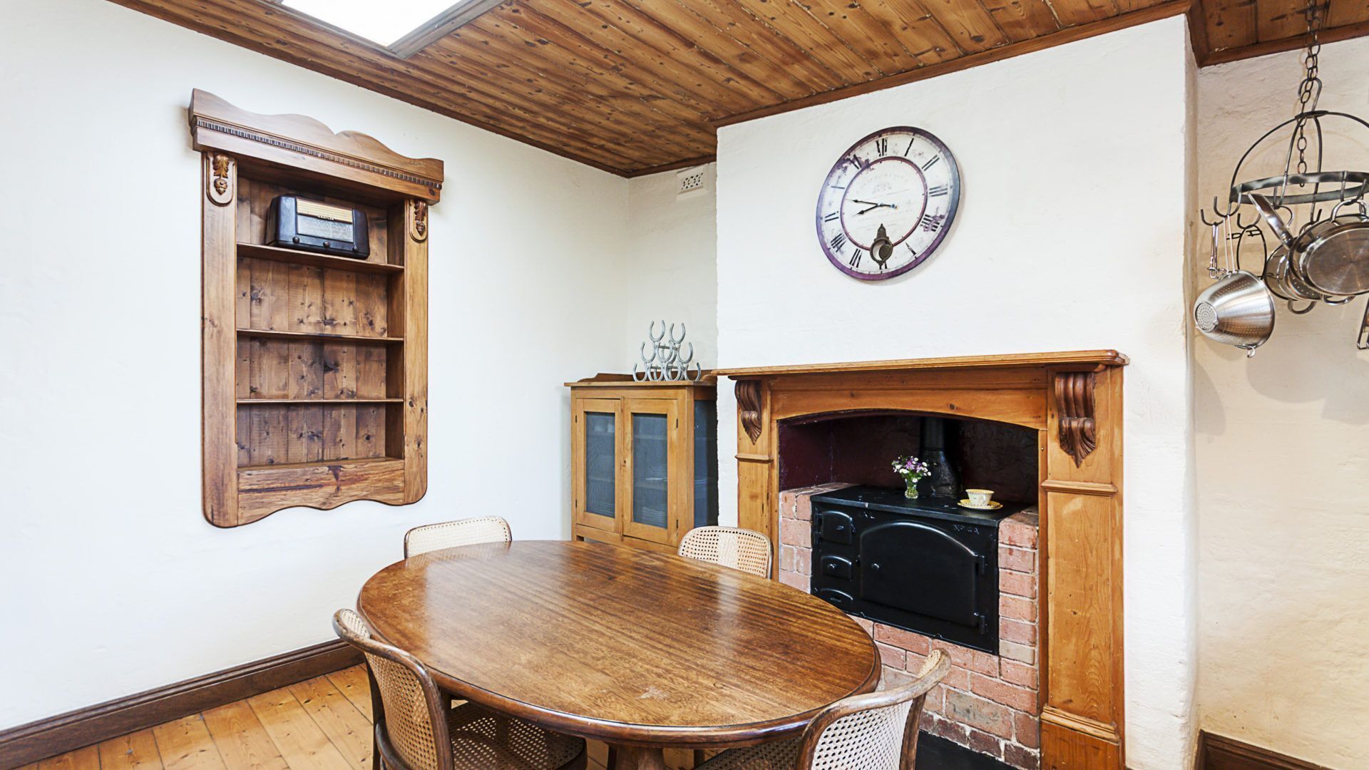 Een eethoek met een houten tafel en stoelen, een houten kast met planken, een klok aan de muur, en een open haard met een metselwerk omlijsting.