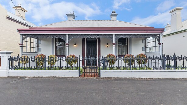 A two-story house with a red-tiled roof, a front porch with decorative iron railings, and a small front yard with potted plants. - Positioned in the center of Warrnambool at a location that cannot be beat. (WARRNAMBOOL)