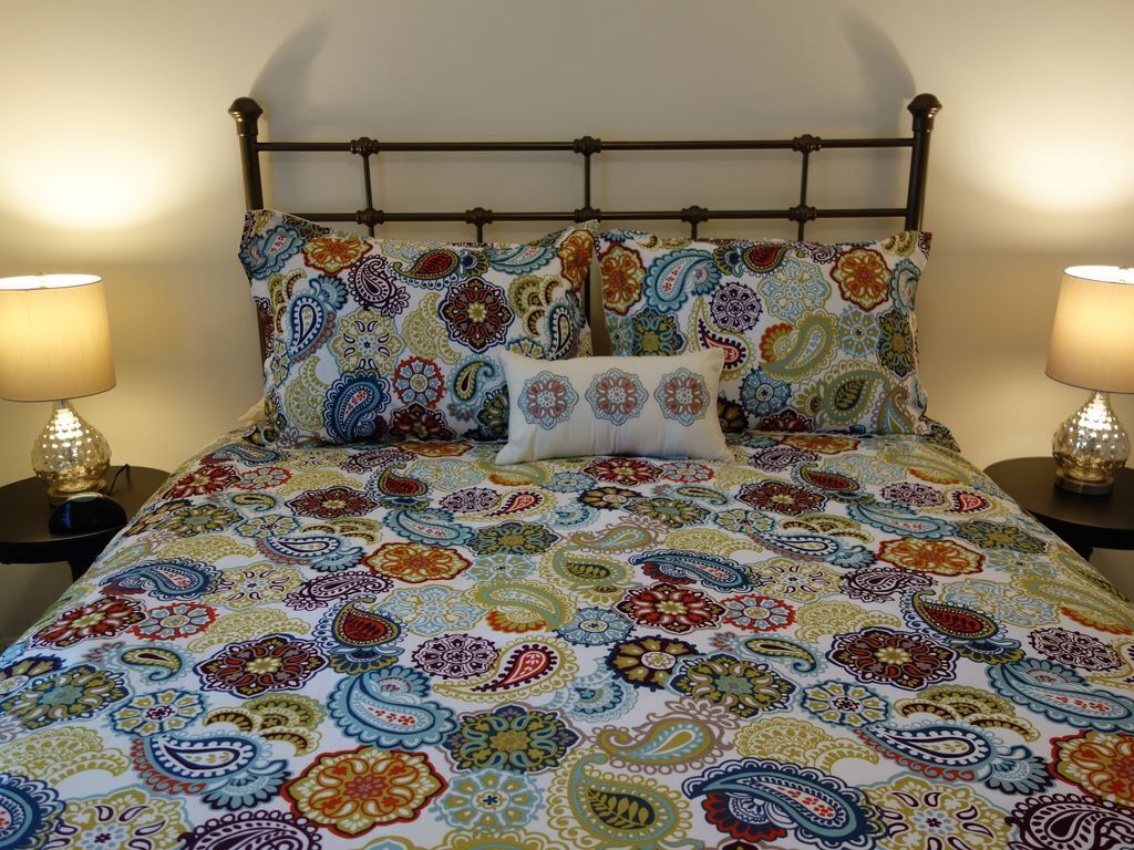 Uma cama com um edredom colorido estampado com padrão paisley, duas luminárias de cabeceira e um abajur decorativo na parede.
