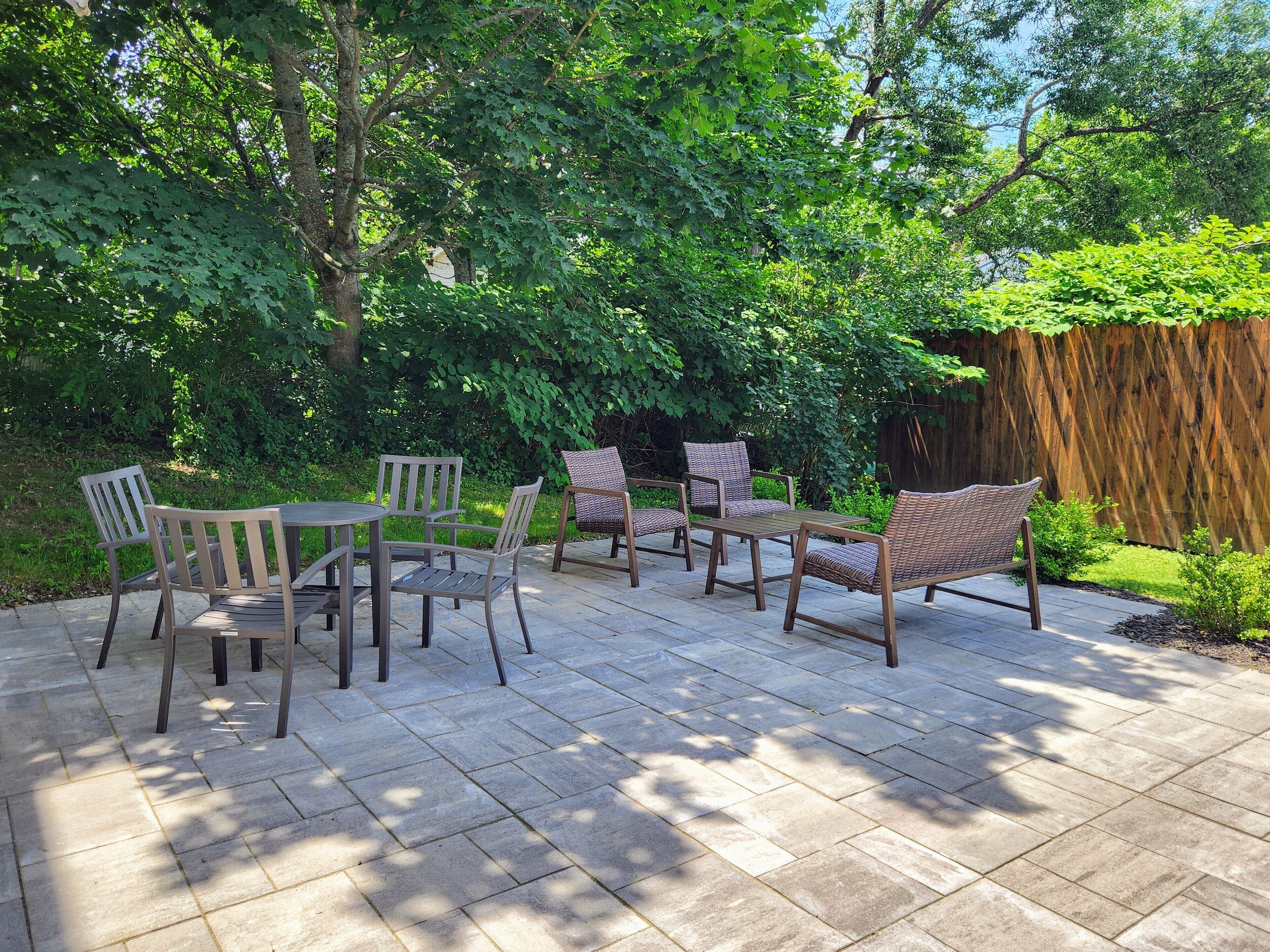Een terras buiten met een tafel en zes rieten stoelen, omringd door weelderige begroeiing en een houten hek.