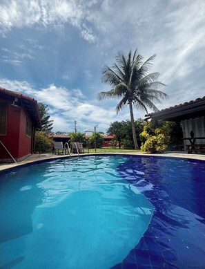 Uma piscina com água azul cristalina, rodeada por um edifício vermelho, uma palmeira e uma área de estar coberta.