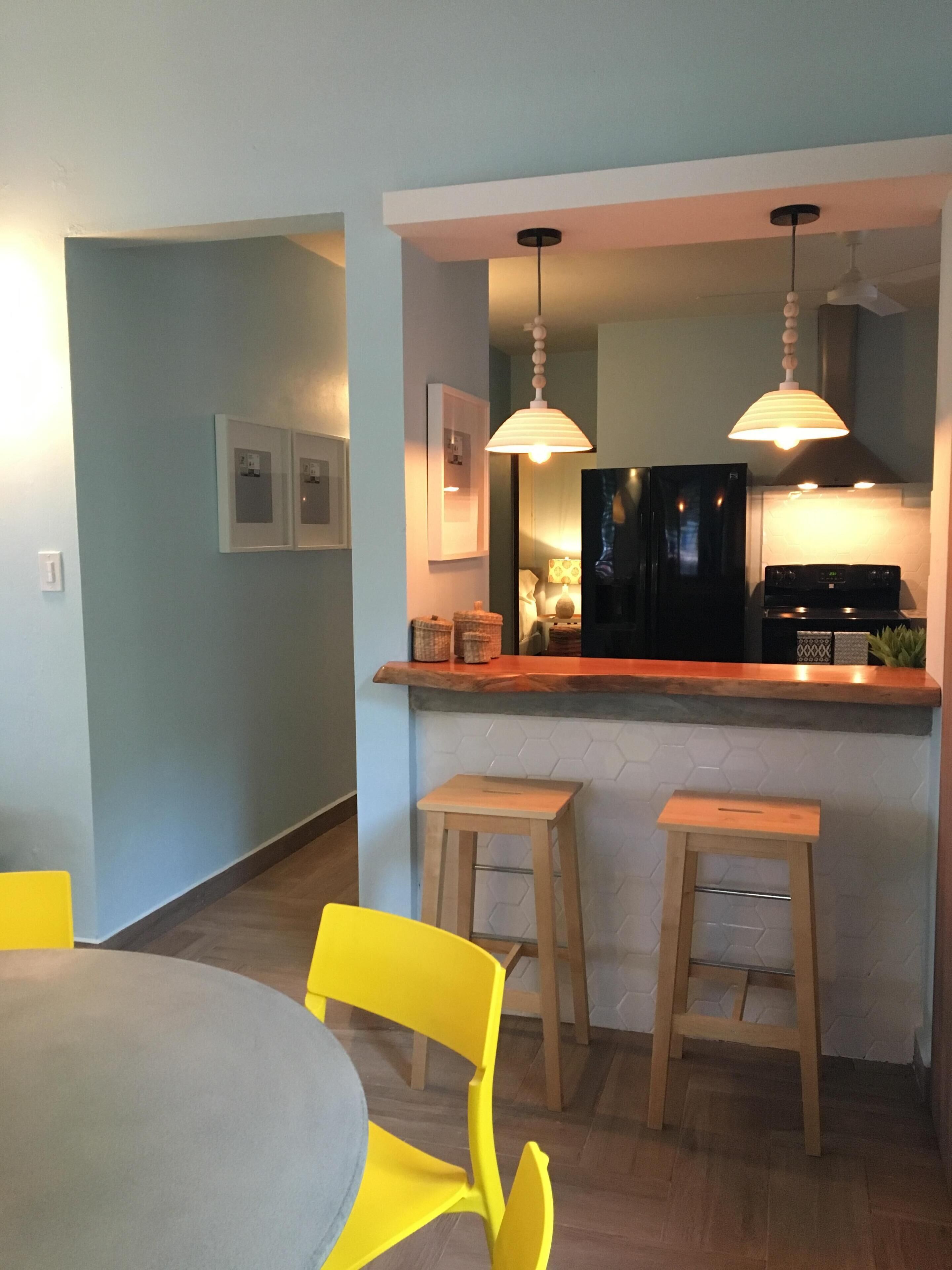 A modern kitchen with a bar area, wooden stools, and a dining table with yellow chairs.